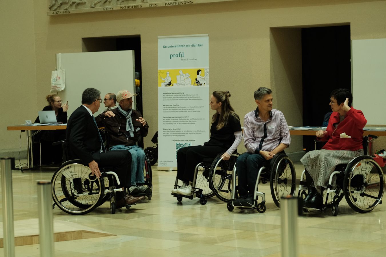 Rollstuhlsimulation: Mehrere Personen im Rollstuhl, darunter die Prorektorin Lehre und Studium, Gabriele Siegert, und der Rektor der UZH, Michael Schaepman, sitzen in einer Runde und unterhalten sich im Lichthof der UZH, mit Informationsstand im Hintergrund.